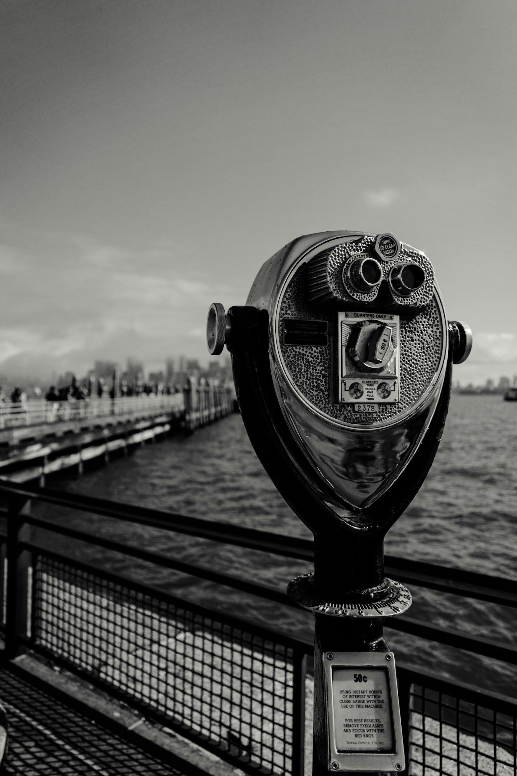 Black and white image of a coin-operated viewer on a pier overlooking the water.